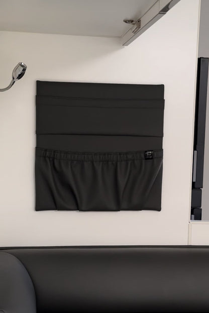 A black viva leatherette fabric wall-mounted caravan storage organizer with two pockets and an elastic in the top of the pocket, shown hanging on a white wall inside a caravan and or motorhome with a dark leather seat visible in the foreground.