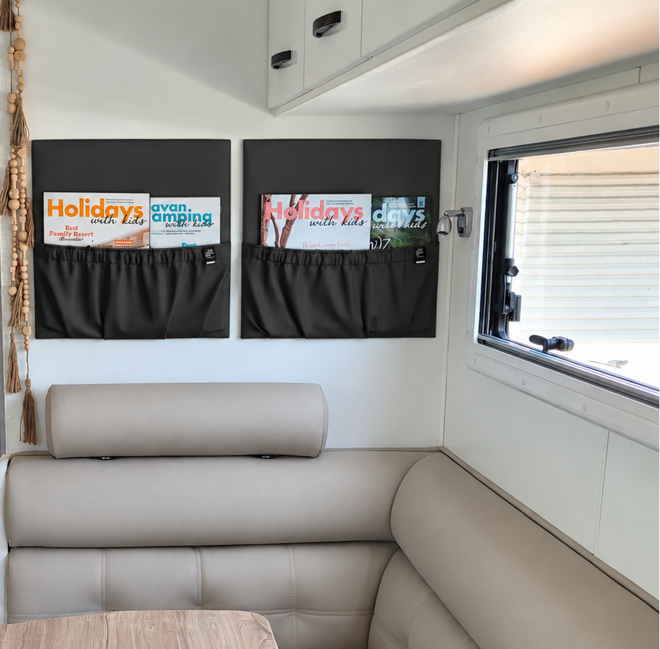 A compact recreational vehicle interior featuring a light beige L-shaped bench seat, a light wood dining table, white cabinets, a washing machine, and two square single black magazine holders on the caravan wall.