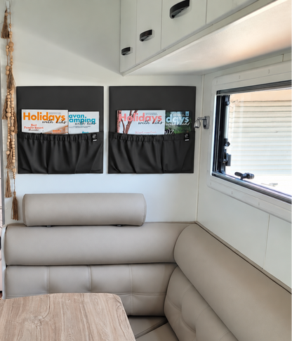 A compact recreational vehicle interior featuring a light beige L-shaped bench seat, a light wood dining table, white cabinets, a washing machine, and two black magazine holders on the wall.